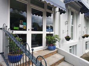 A building with a white facade, a blue awning, and a 'NO VACANCIES' sign. There are potted plants on the steps and hanging flower baskets. - Harmony Guest House (Torquay)