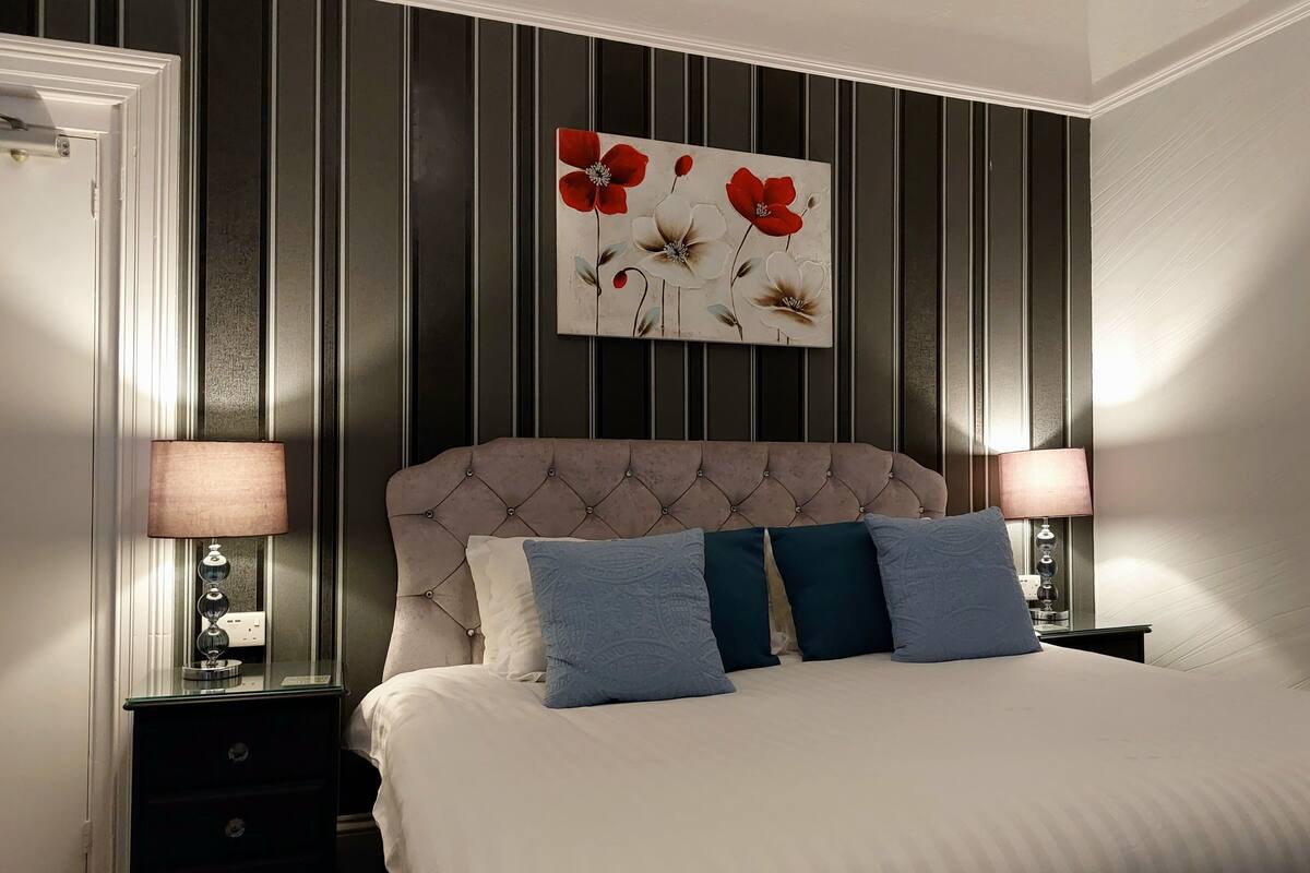 A bedroom with a tufted headboard, a large bed with white and blue bedding, two bedside tables with lamps, and a floral artwork above the bed.