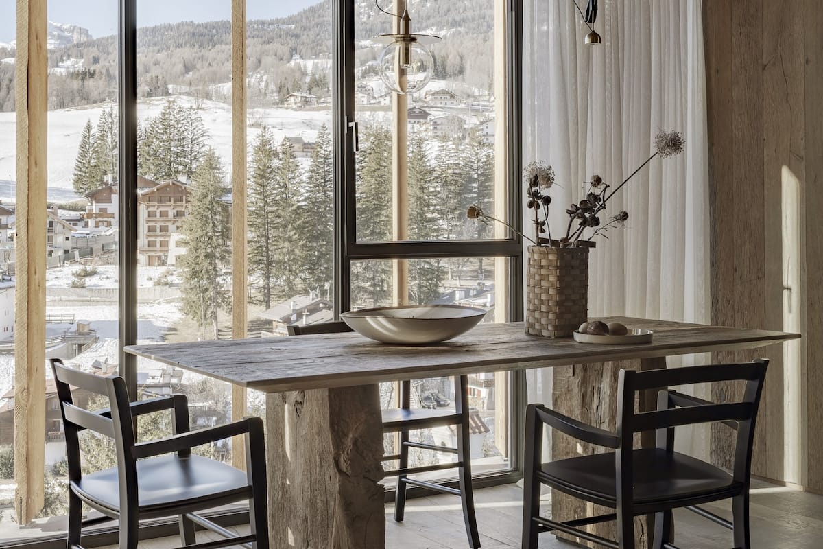 En matplats med ett stort träbord, två stolar, en vas med torkade blommor och utsikt över ett snötäckt landskap genom stora fönster.