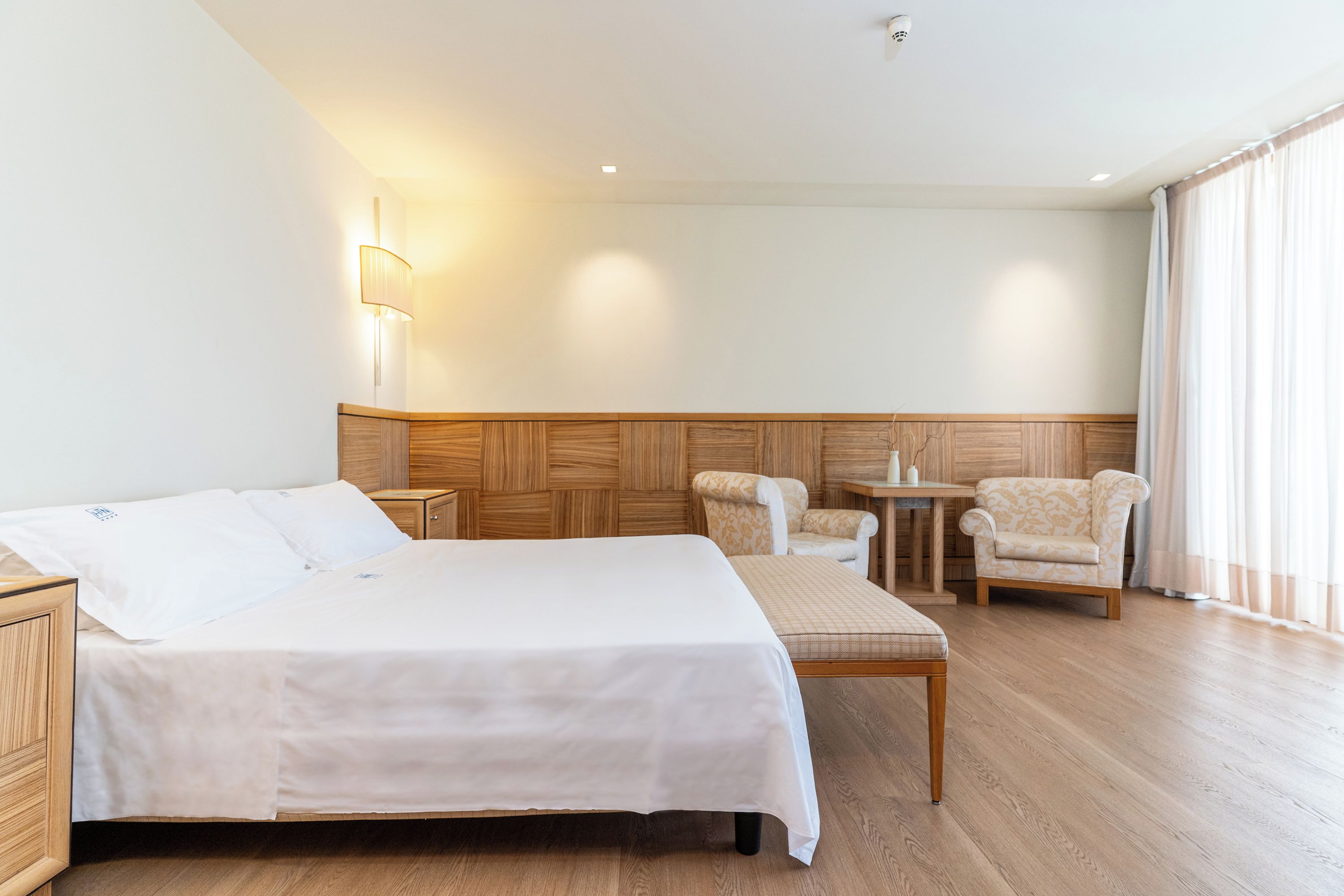 Una camera d'albergo con un letto grande, due poltrone, un tavolino e una testiera in legno.