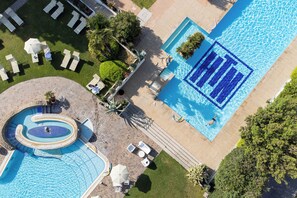 Vista aérea de um resort com uma piscina grande, uma piscina circular menor e várias áreas de descanso com guarda-sóis.