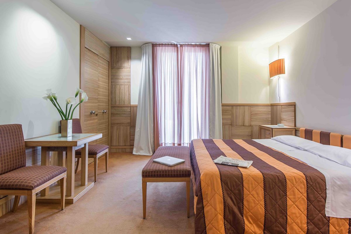 A hotel room with a bed, a desk, a chair, a lamp, and a window with curtains.