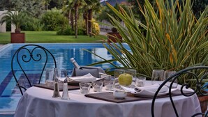 Une table à manger dressée avec une nappe blanche, des verres, des couverts et une bouteille, donnant sur une piscine bordée de plantes tropicales.
