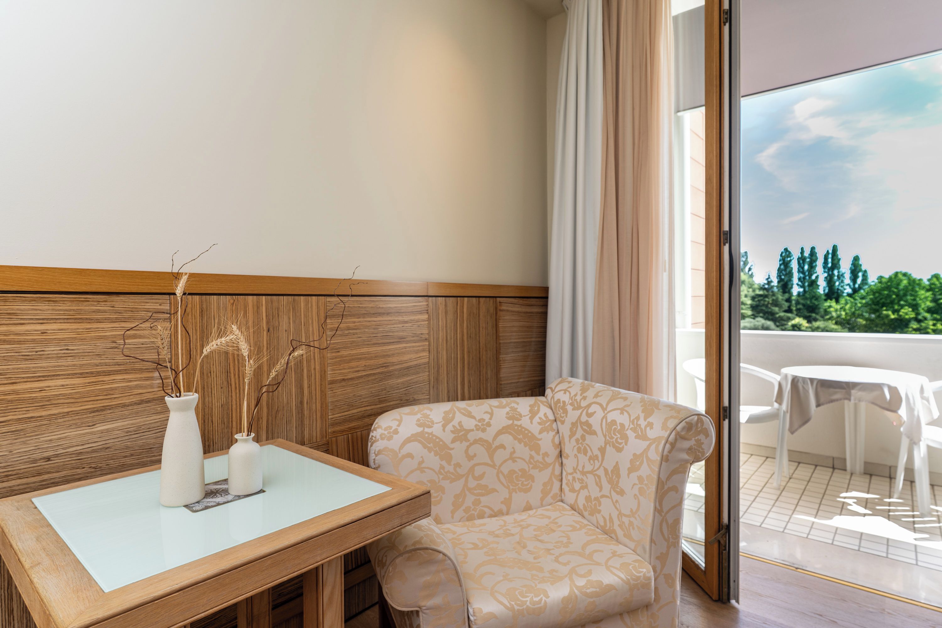 A room with a wooden headboard, a white table with two vases, a patterned armchair, and a view of greenery through a glass door.