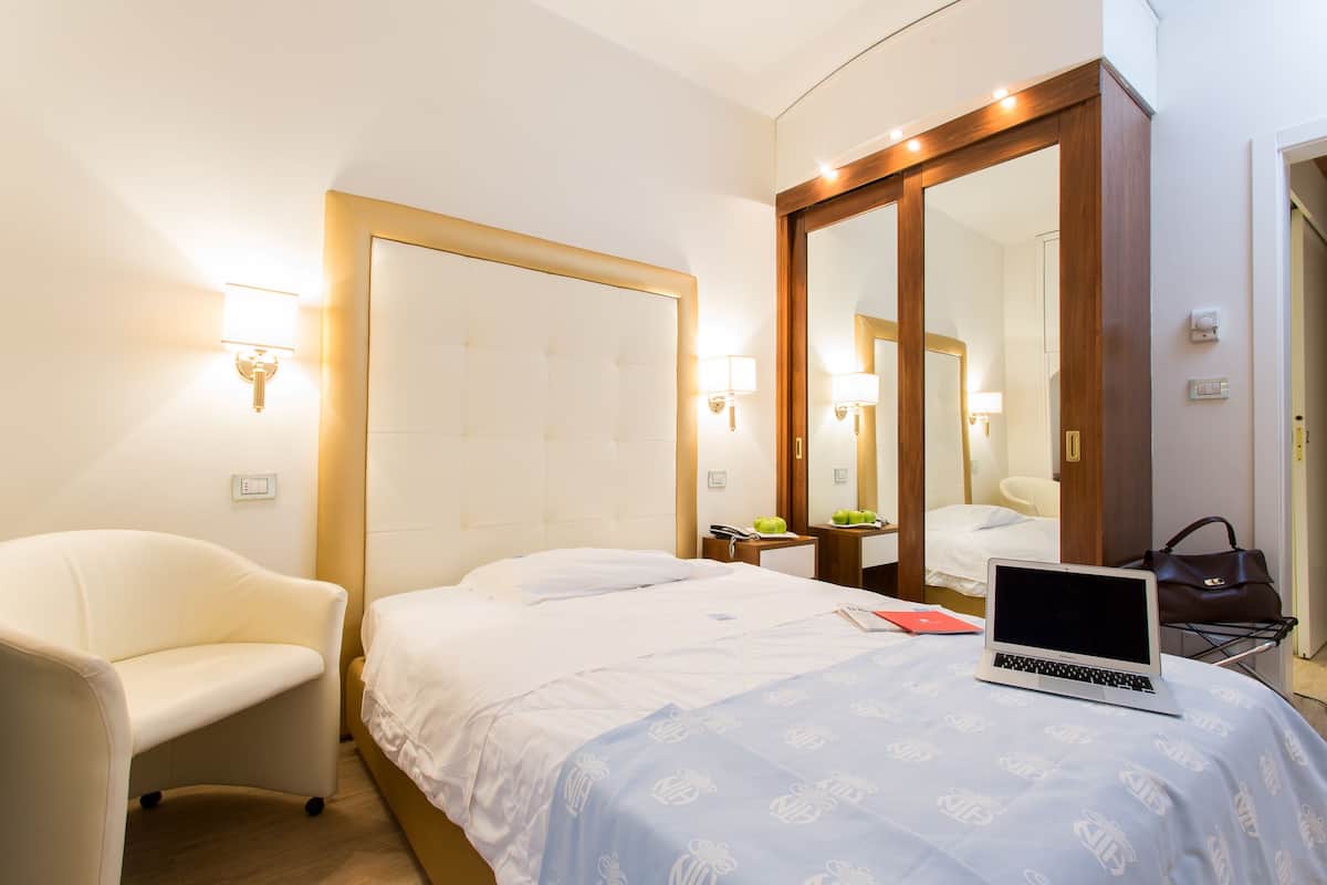Ein Hotelzimmer mit einem Bett, einem Sessel, einem Schreibtisch mit einem Laptop, einem Spiegel und einer Handtasche.