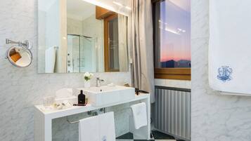 A modern bathroom with a large mirror, two sinks, a shower, and a view of the sunset through a window.