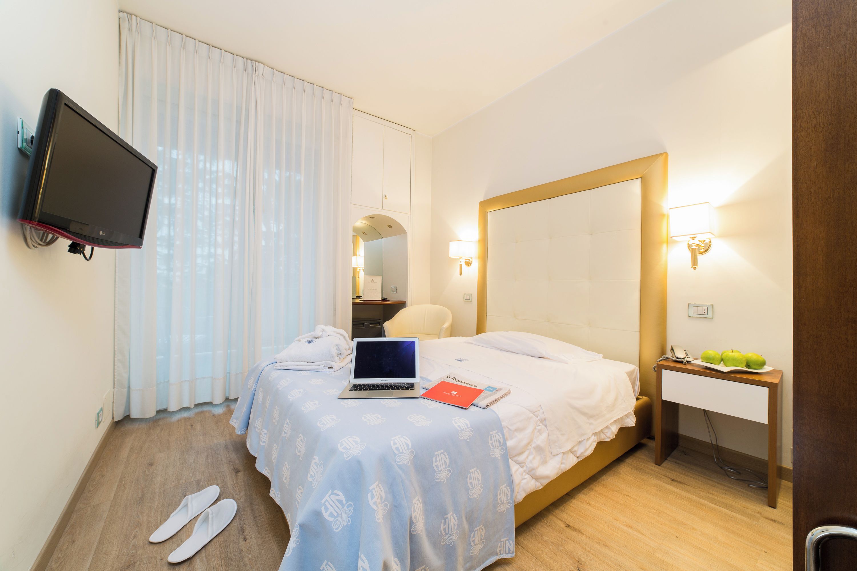 Habitación de hotel con cama, escritorio con una computadora portátil, televisor y mesita de noche con lámpara.