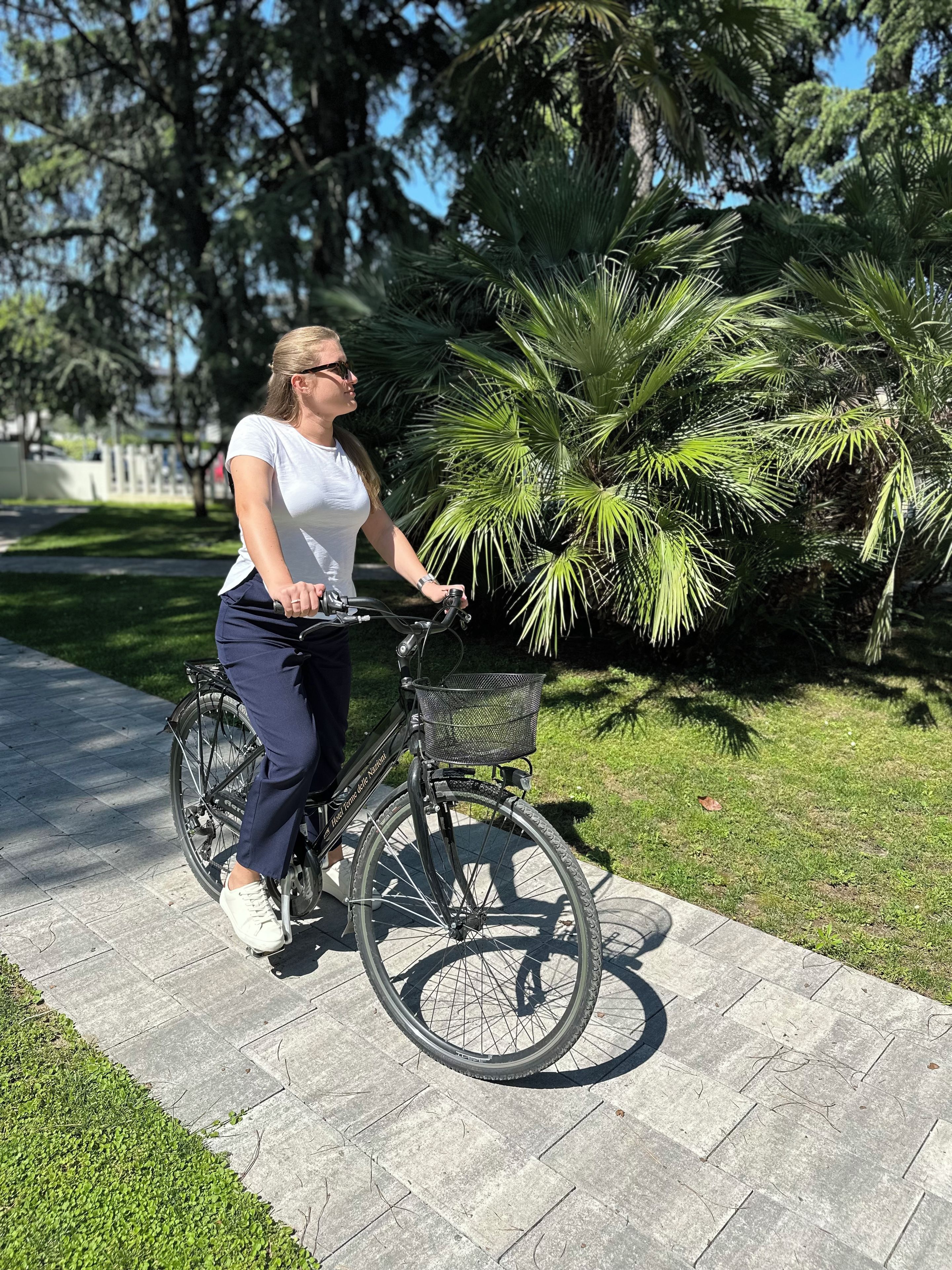 A person on a bicycle with a basket, parked on a paved path with grass and trees in the background.