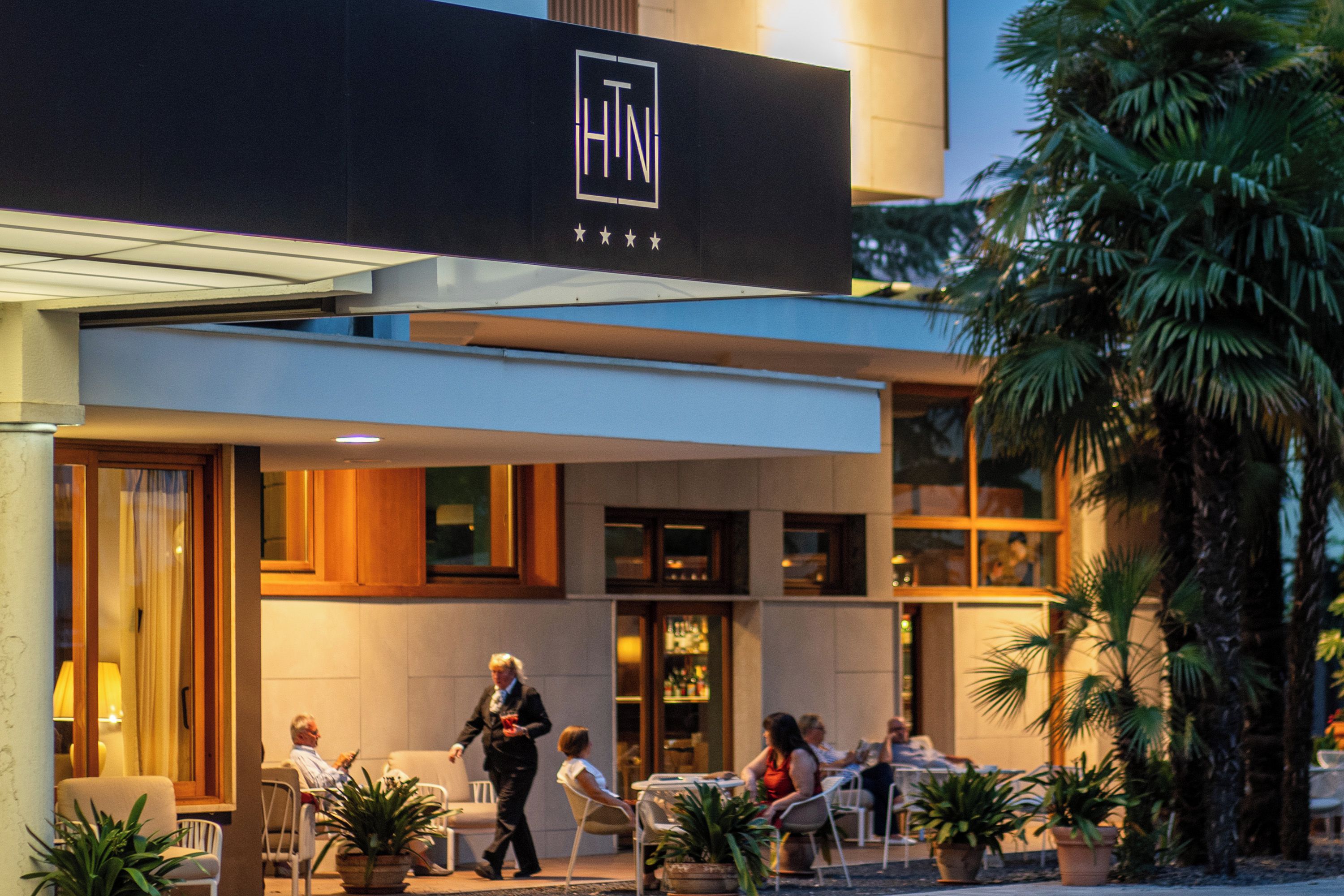 En hotellfasad med uteservering, palmer och en skylt med logotypen ”HTN”.