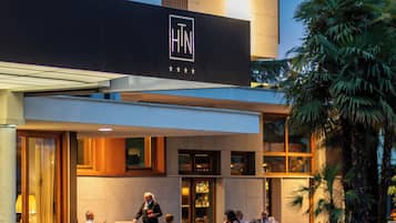 En hotellfasad med uteservering, palmer och en skylt med logotypen ”HTN”.