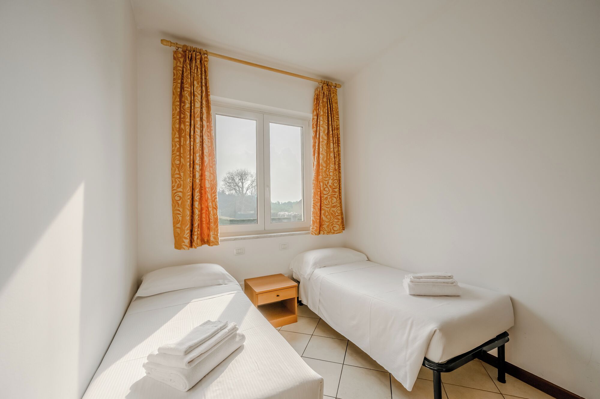 A small, clean room with two single beds, a window with curtains, and a wooden nightstand.