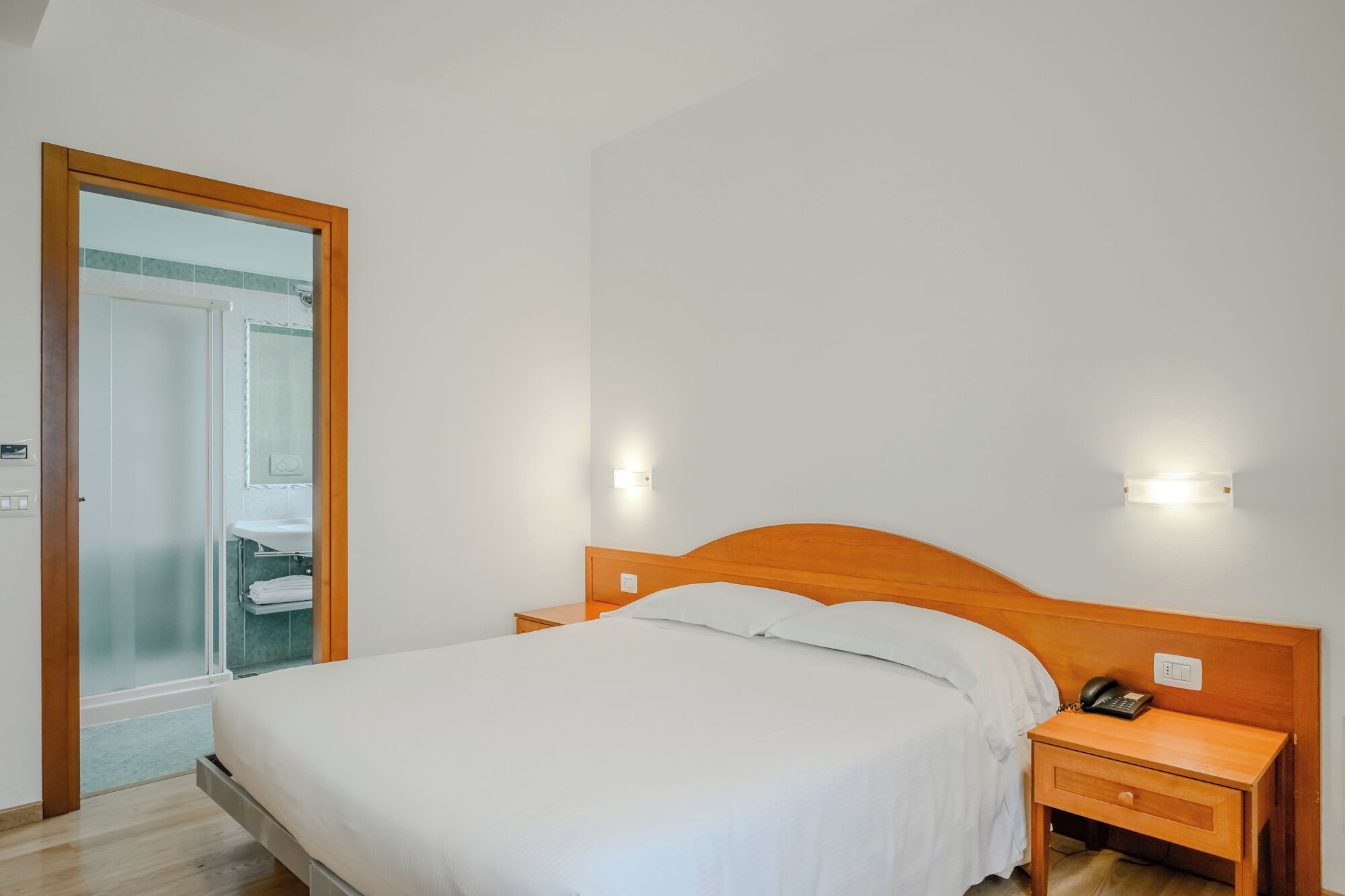 A hotel room with a bed, bedside table, and a bathroom visible through a glass door.