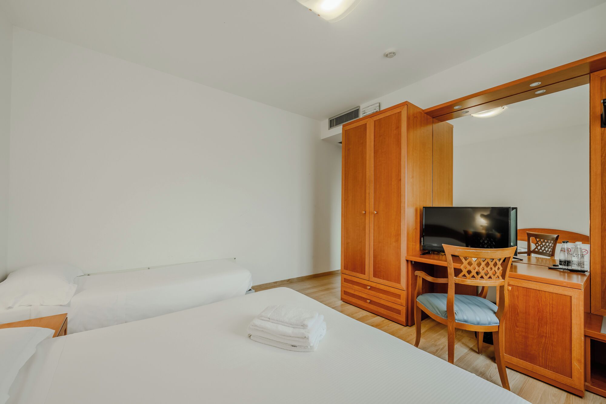 A hotel room with a bed, a TV, a wooden wardrobe, and a desk with a chair.