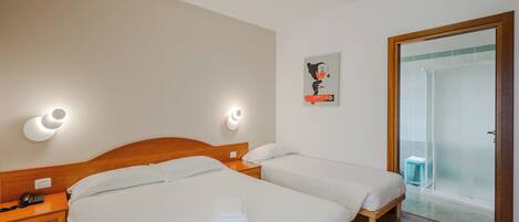 A hotel room with two beds, a wooden headboard, bedside tables with lamps, a telephone, a framed picture on the wall, and a sliding glass door leading to a shower area.