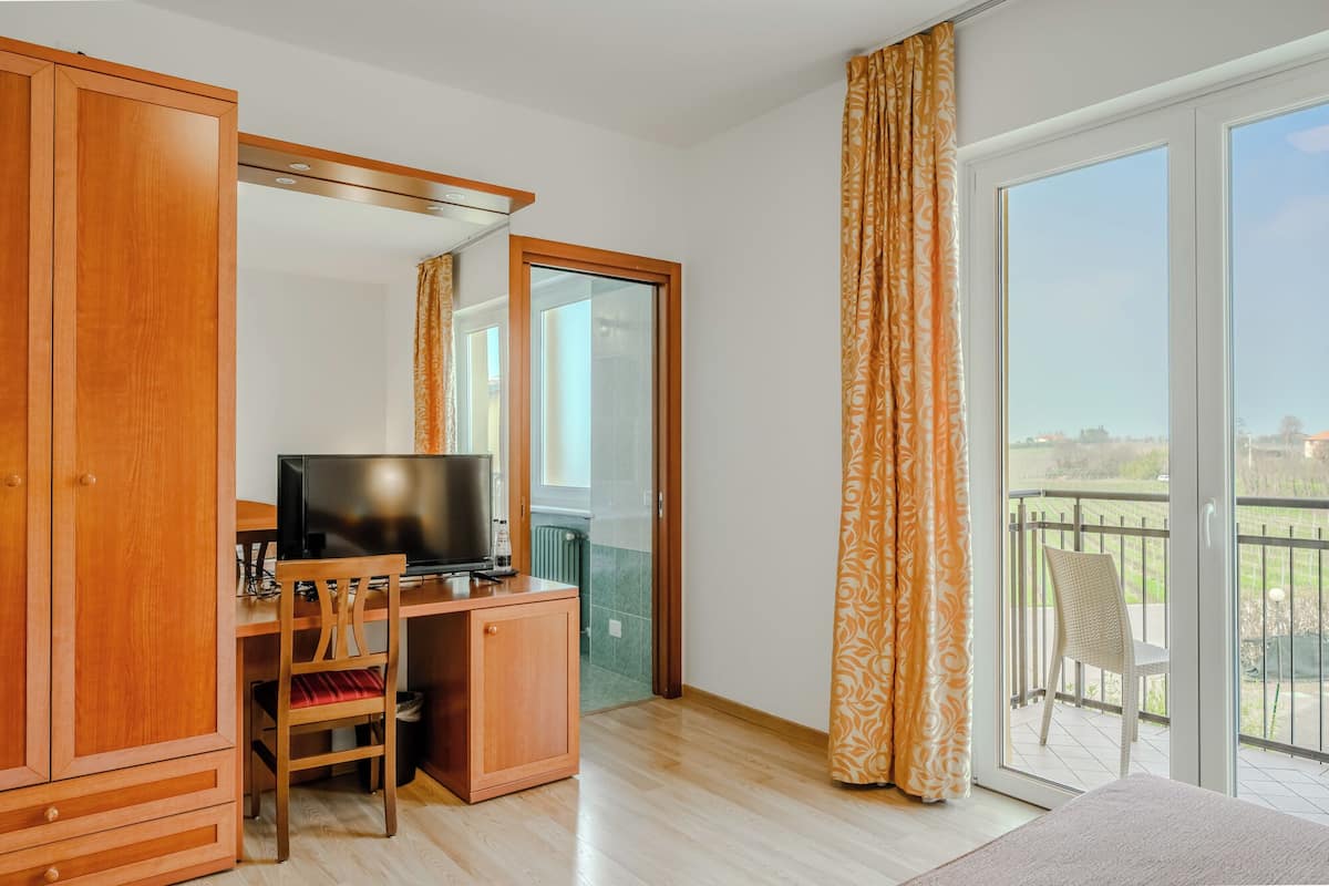 Ein Zimmer mit einem HolztV-Schrank, einem Flachbildfernseher, einem kleinen Tisch und einem Sessel. Eine Glastür öffnet sich zu einem Balkon mit Blick auf die Landschaft.