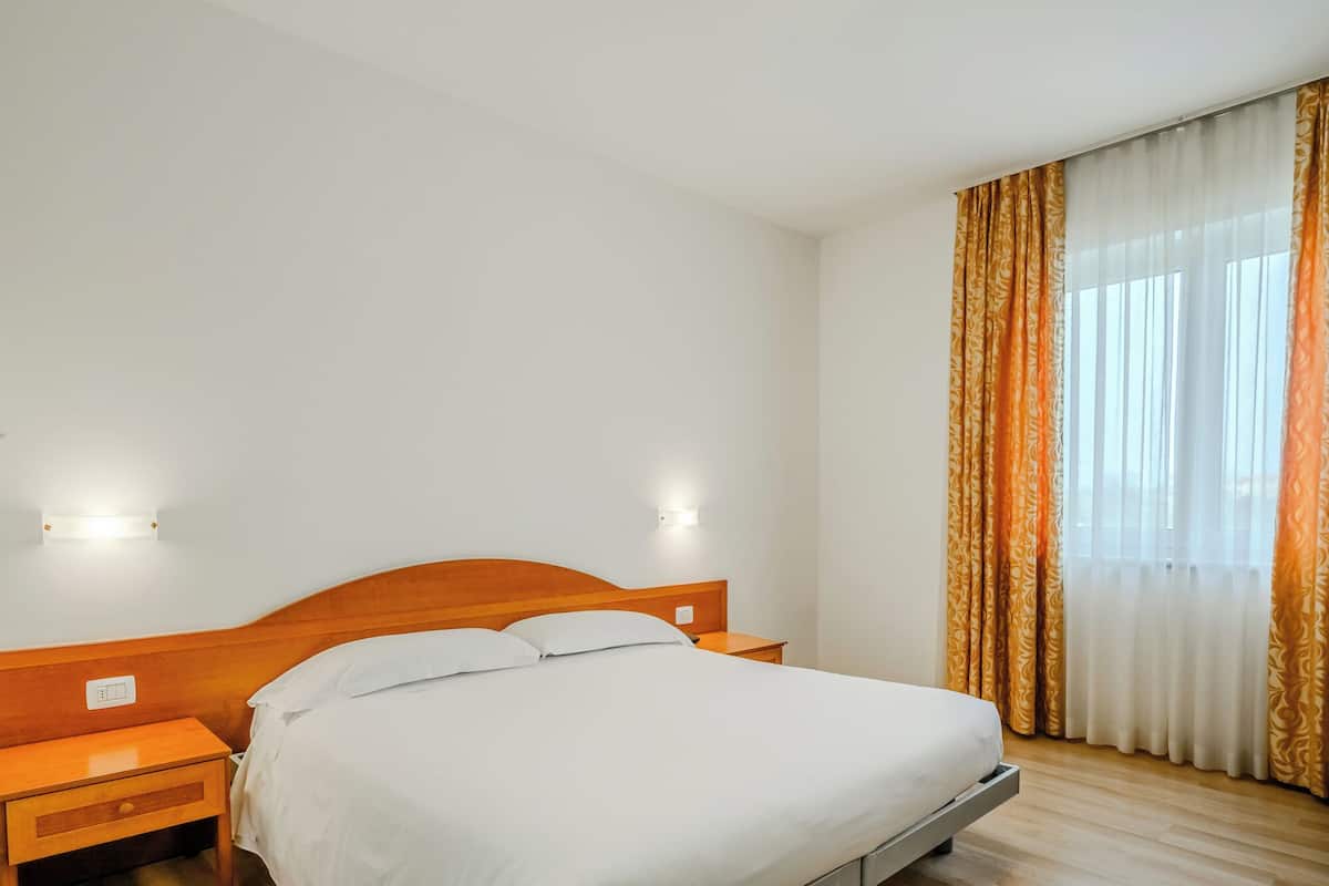 Ein Hotelzimmer mit hölzernem Kopfteil, einem Einzelbett, einem Nachttisch und einem Fenster mit Vorhängen.