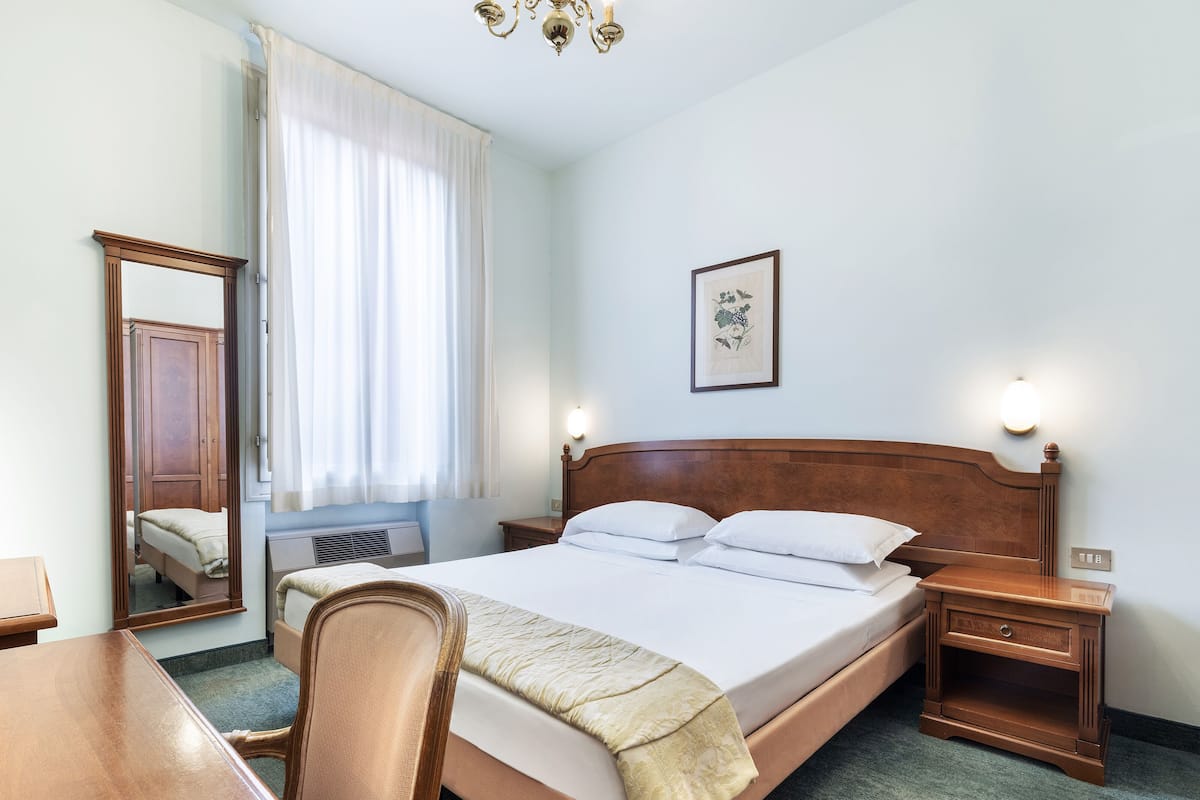 A hotel room with a wooden bed, a desk, a chair, a mirror, a window with curtains, a wall-mounted light, and a framed picture.