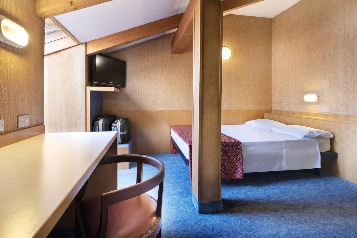 A compact room with a bed, a desk, and a chair. There is a television mounted on the wall and a small table with a chair. The room has wooden beams on the ceiling and a carpeted floor.