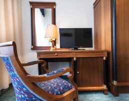A wooden chair with a patterned cushion, a desk with a television, and a mirror on the wall.