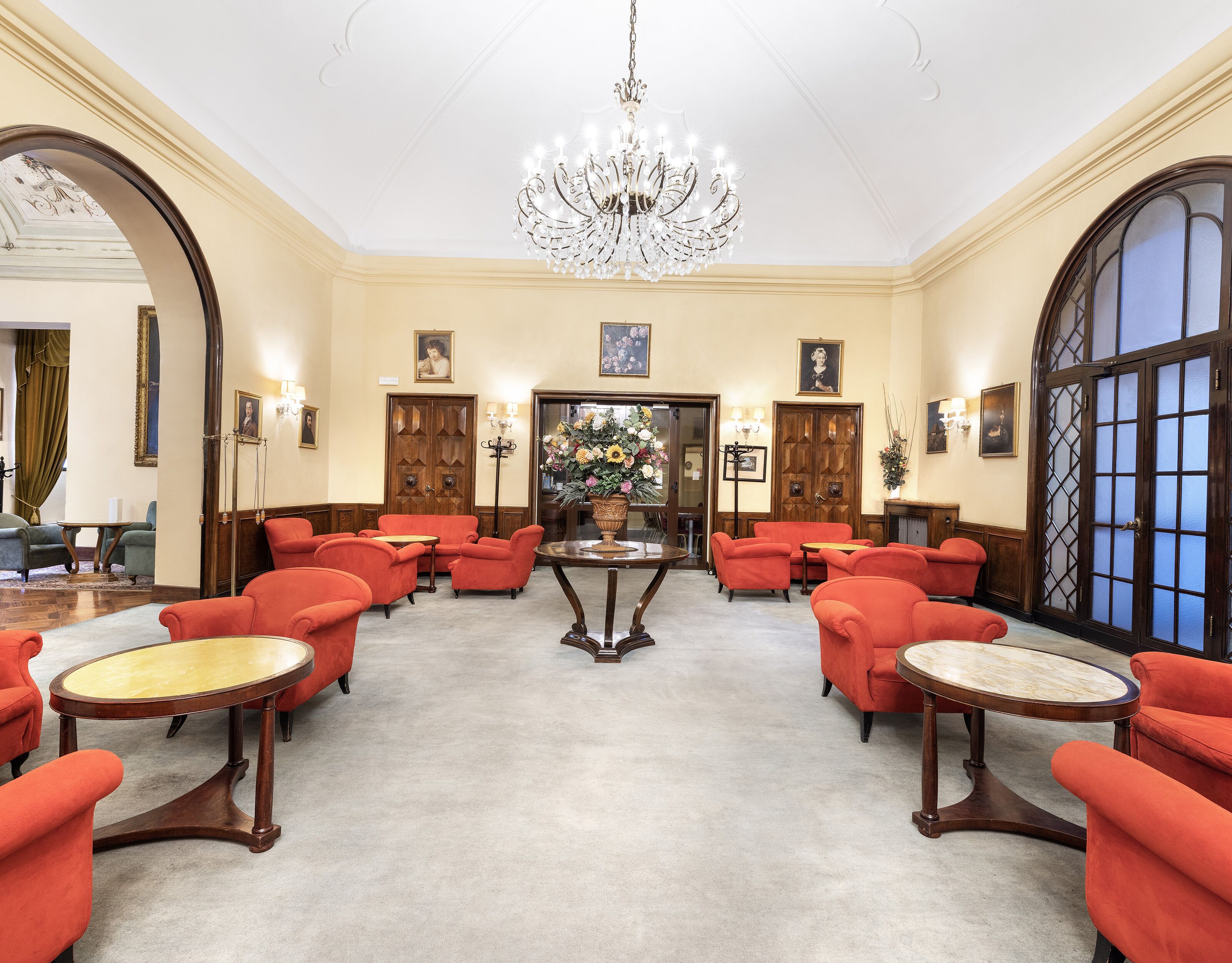 A spacious lobby with a grand chandelier, red upholstered chairs, and a central table.