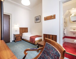 Een hotelkamer met een bed, nachtkastje, stoel en een badkamer met wastafel en spiegel.