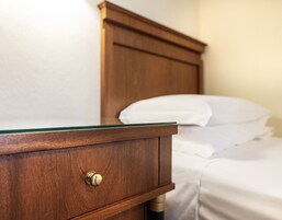 Un comodino in legno con un cassetto e una maniglia in ottone, un cuscino bianco su un letto e una testiera in legno.