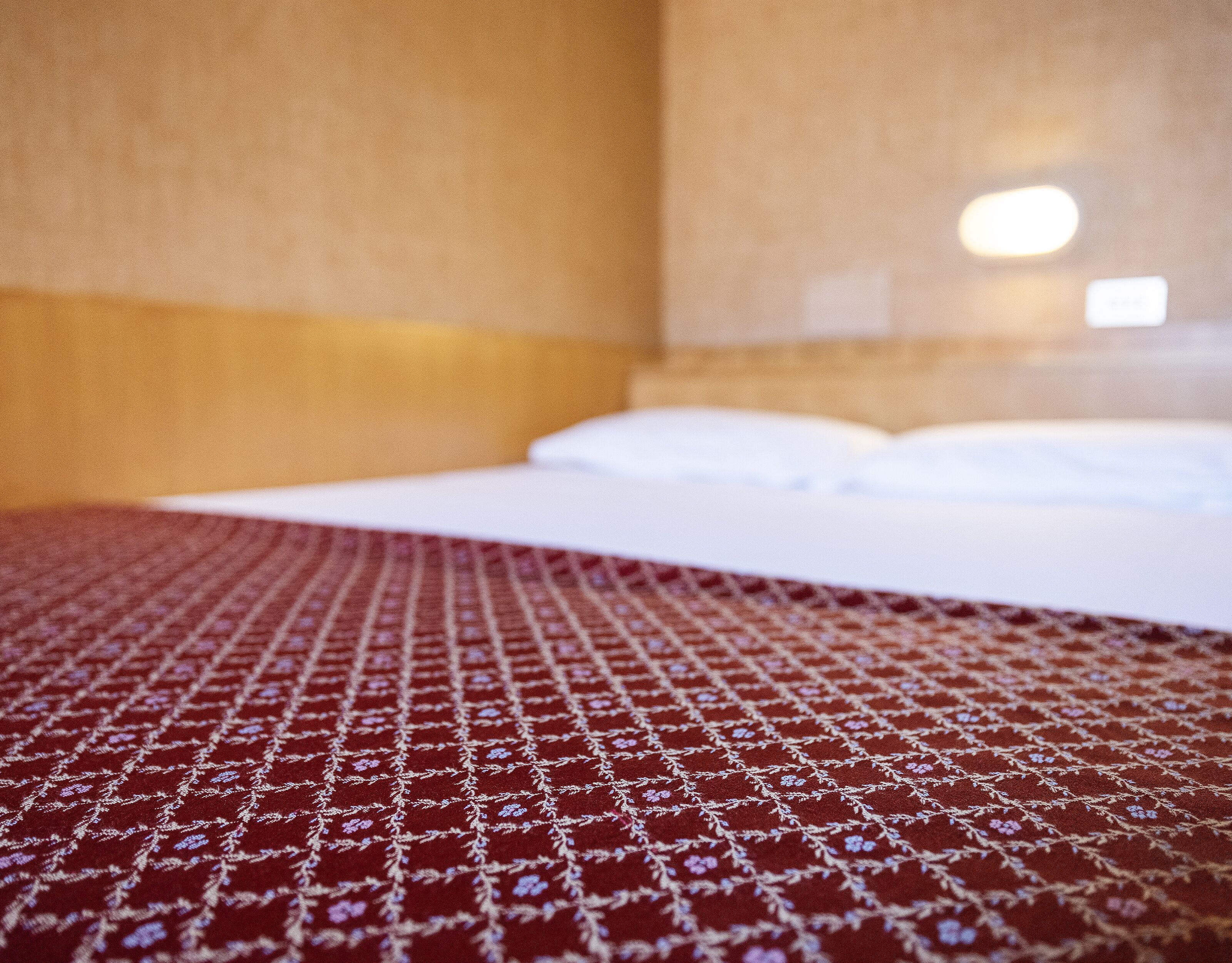 Een hotelkamer met een net opgemaakt bed, een beddengoed met een rood-wit patroon, en een effen muur met een lichtschakelaar.