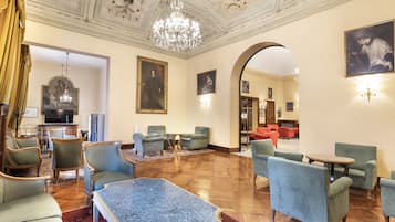 Een ruime lobby met een hoge plafond en een gedetailleerd plafondontwerp, inclusief een kroonluchter. Er zijn diverse zithoeken met fauteuils en een salontafel.