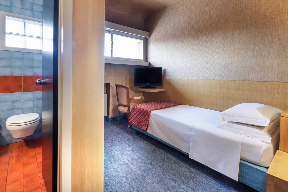 A compact hotel room with a single bed, a small desk with a TV, a chair, and a toilet.