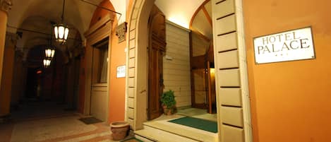 A hotel entrance with a sign reading "HOTEL PALACE," a green mat at the steps, and a hallway with arches and lighting fixtures.