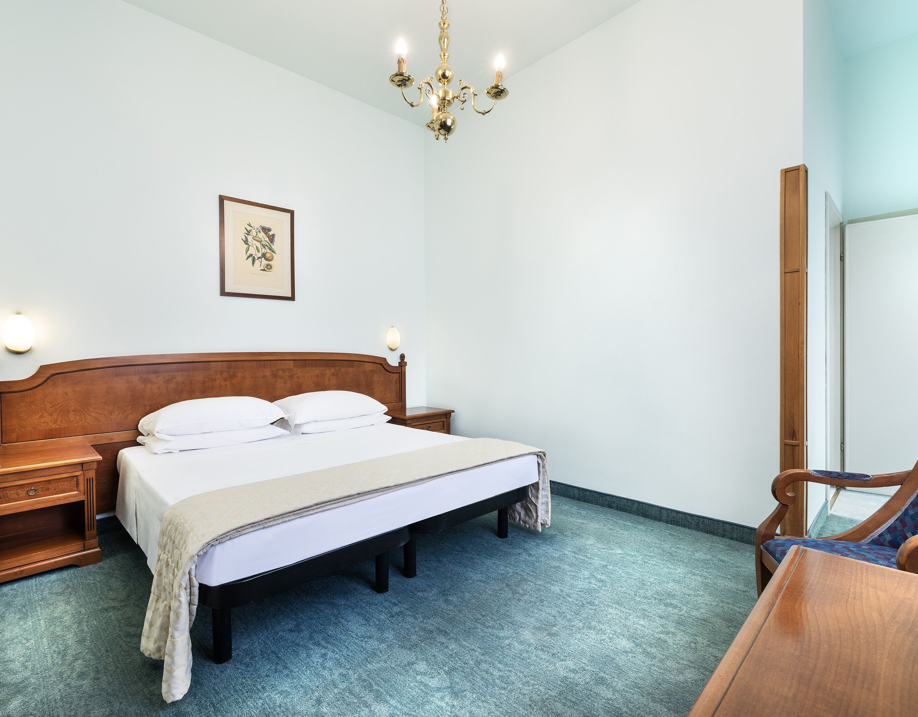 A neatly made bed with a wooden headboard, a bedside table, and a nightstand. There is a framed picture on the wall, a chandelier, and a mirror on the wall.