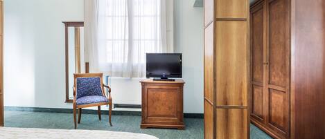 Habitación de hotel con cama, televisión sobre un mueble de madera, una silla, armario y ventana con cortinas.