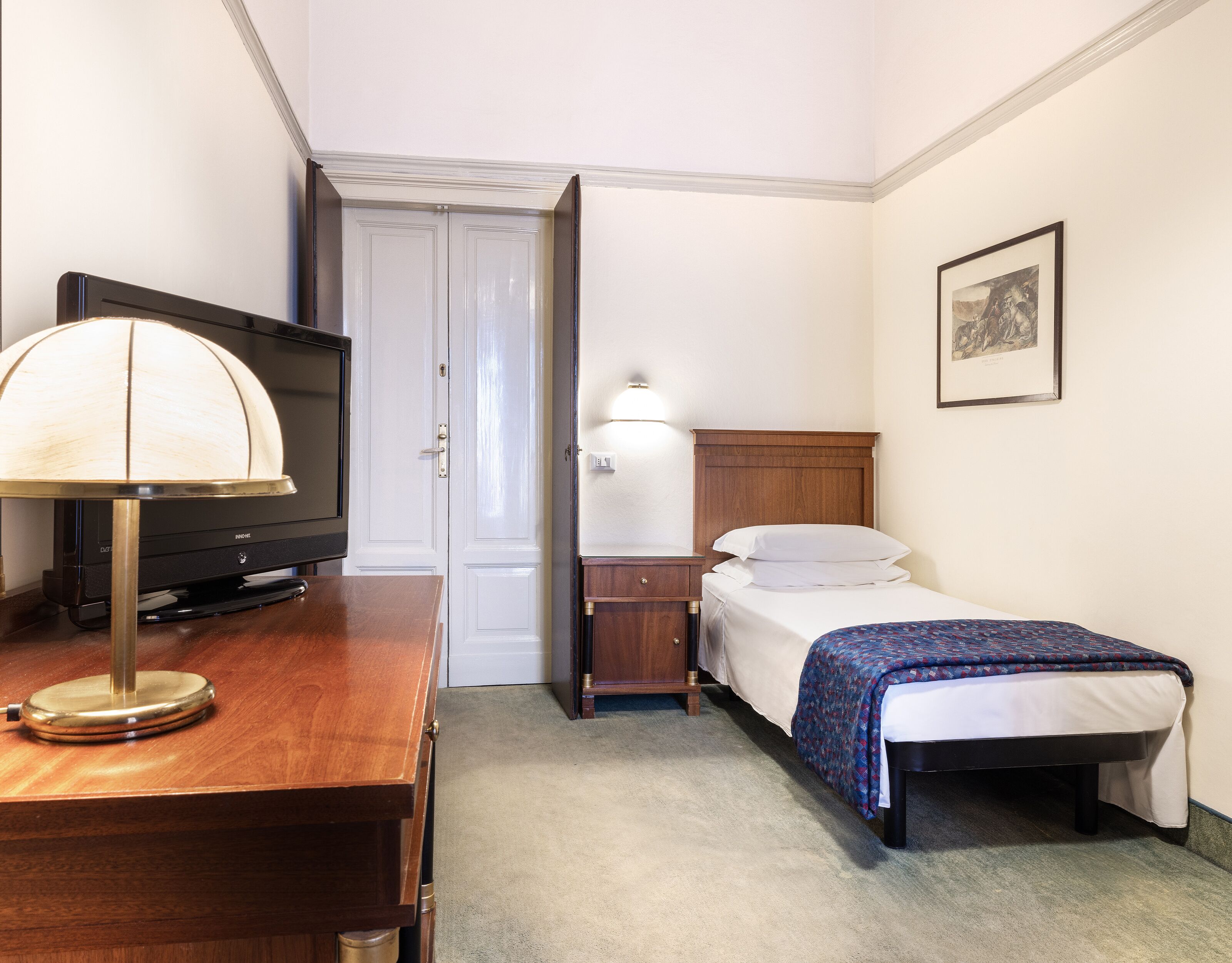 Una camera d'albergo con un letto, un comodino, una lampada, una TV e una porta che dà su un'altra stanza.