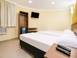 Quarto de hotel com duas camas, televisão presa na parede, minibar e mesa de cabeceira de madeira com dois controles remotos.