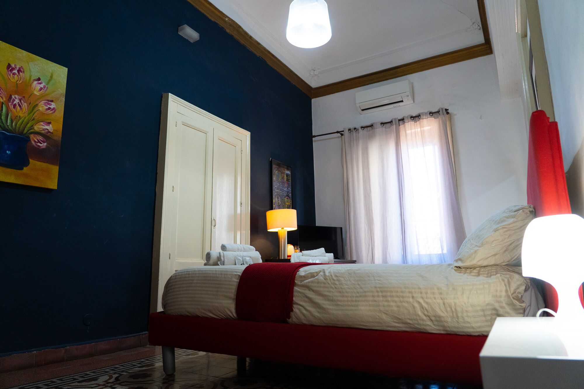 A bedroom with a bed, a nightstand, a lamp, a painting, a wardrobe, and a window with curtains.
