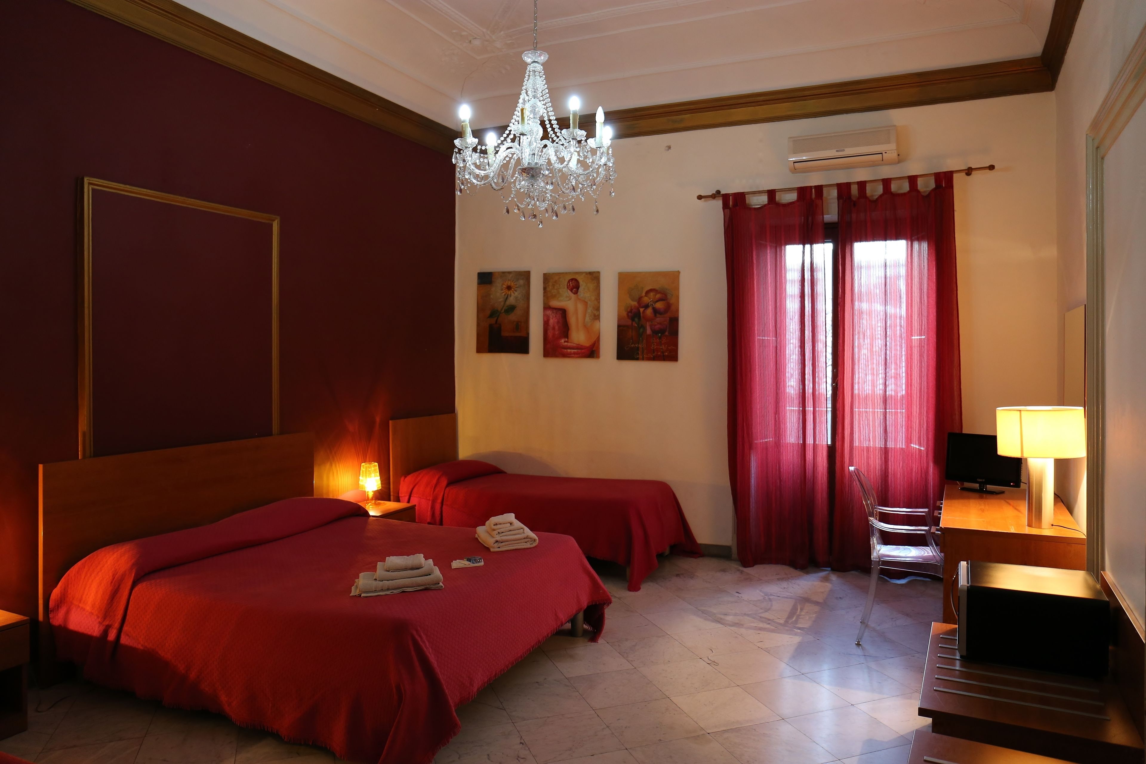 Habitación de hotel con dos camas, un candelabro, un escritorio con lámpara, una silla y obras de arte en la pared.
