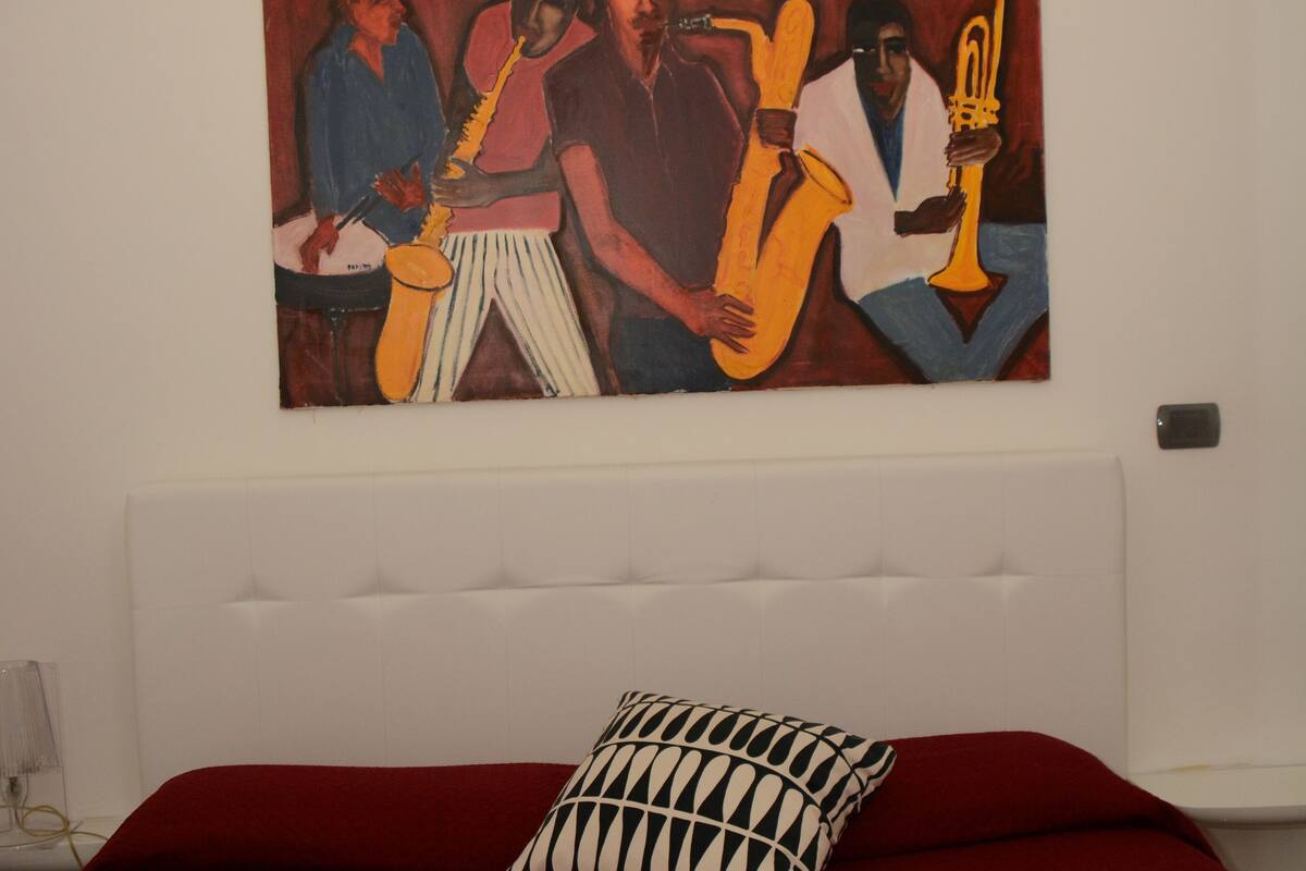 A hotel room with a large painting on the wall, a red bedspread, a white pillow, and a rolled white towel on the bed.