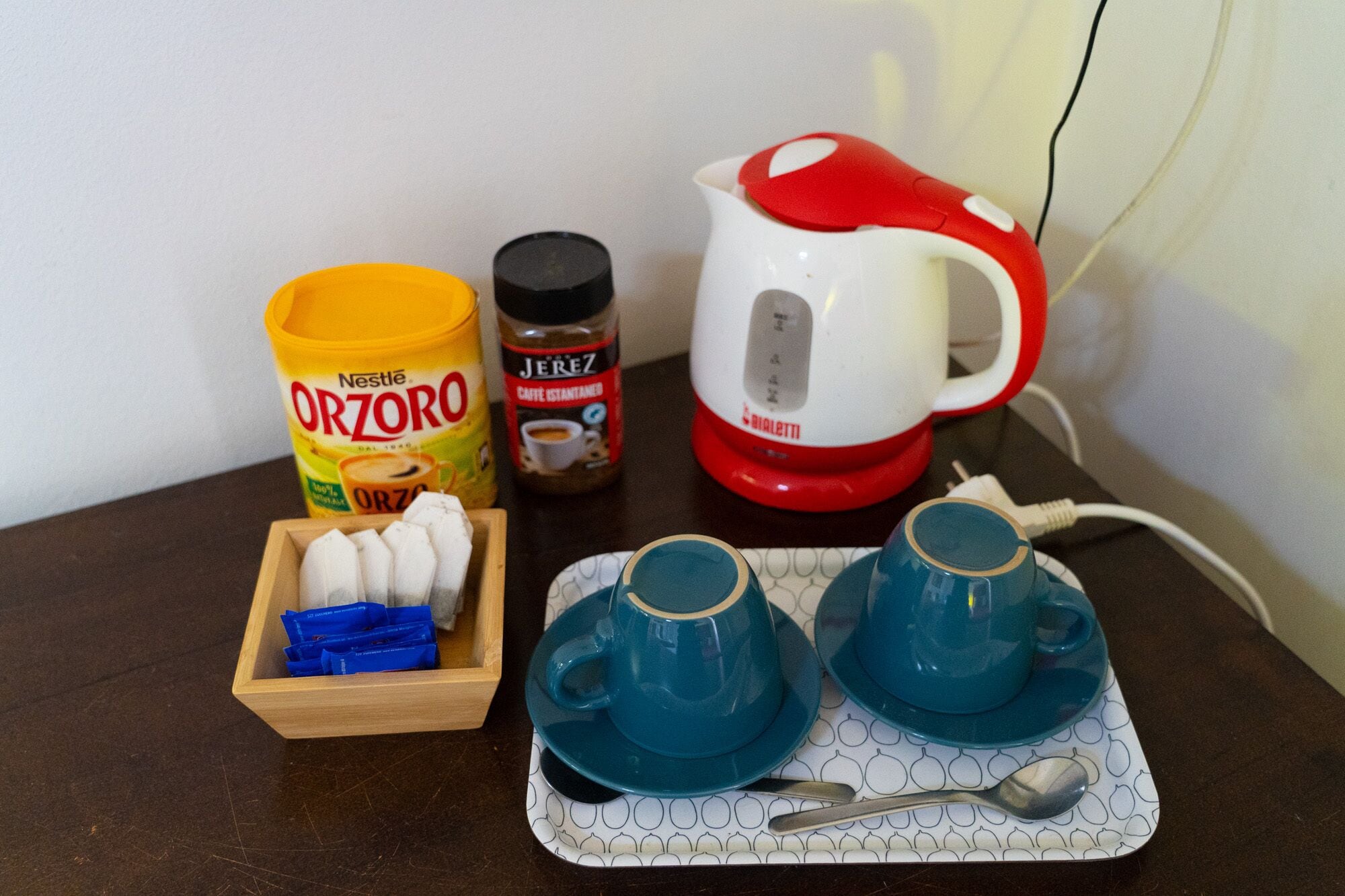 A kitchen counter with a coffee maker, a jar of Nestle Orzoro, a jar of Jerez coffee, a box of blue coffee pods, and two blue cups on a tray.
