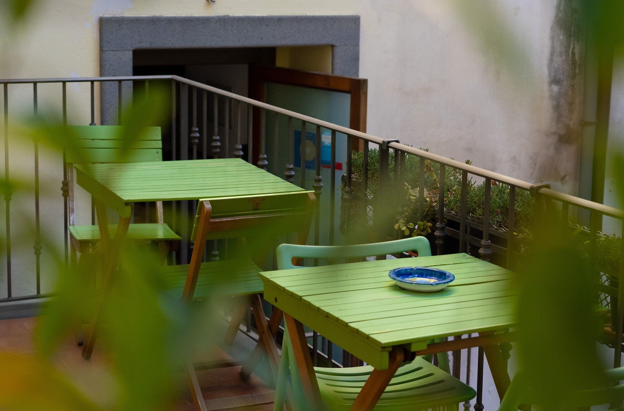 Een balkon met twee groene tafels en stoelen, een blauw bord op één tafel, en een deur die naar een kamer leidt.