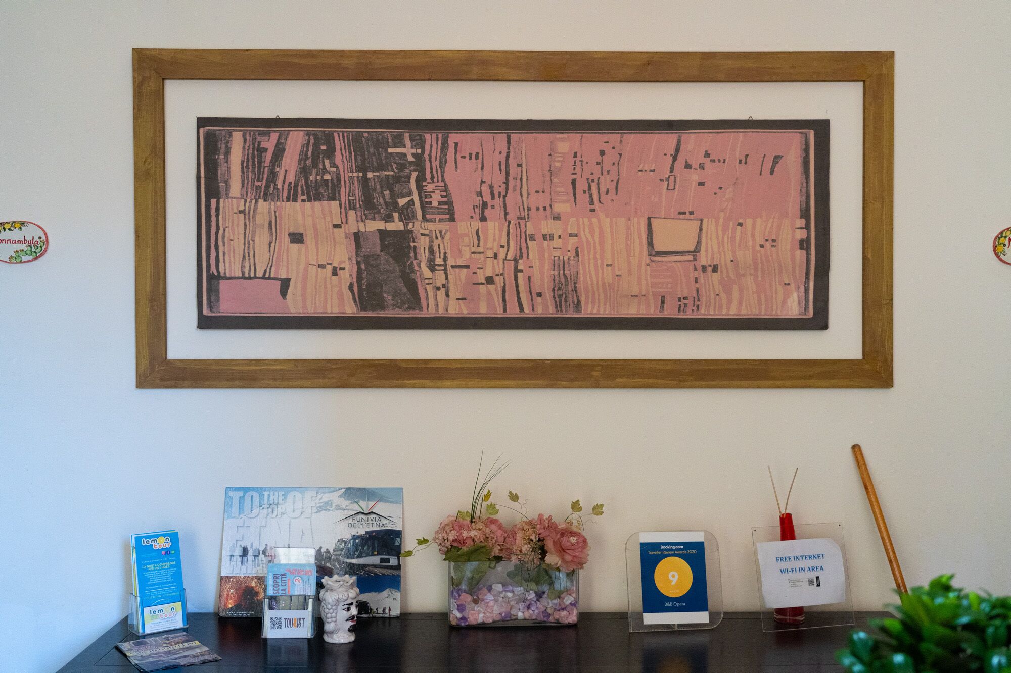 Un cuadro abstracto enmarcado en una pared, un jarrón pequeño con flores, un folleto de viajes, un cartel de Wi-Fi gratuito y un adorno sobre una mesa.