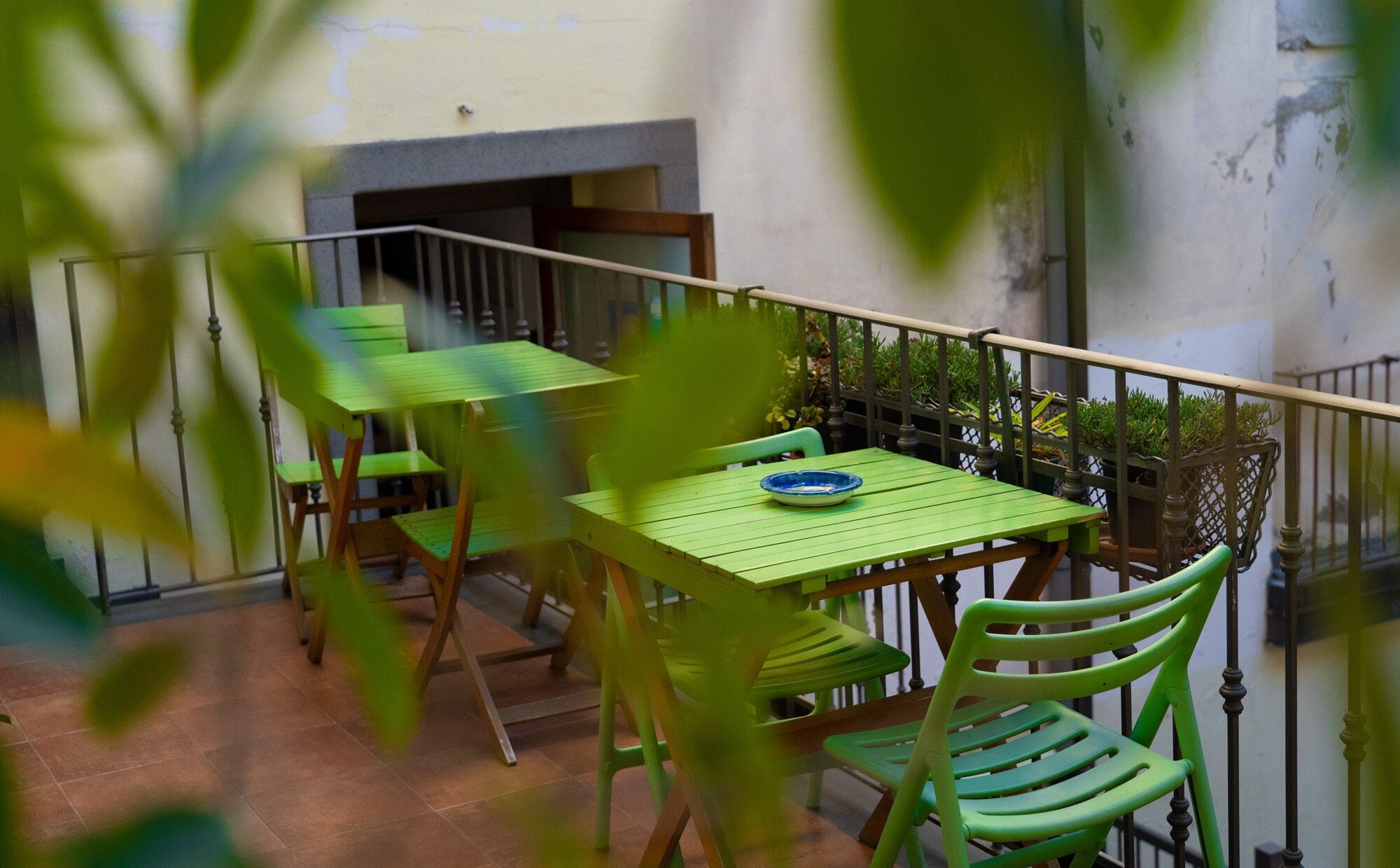 A balcony with green tables and chairs, a blue plate on one table, and a railing with potted plants.