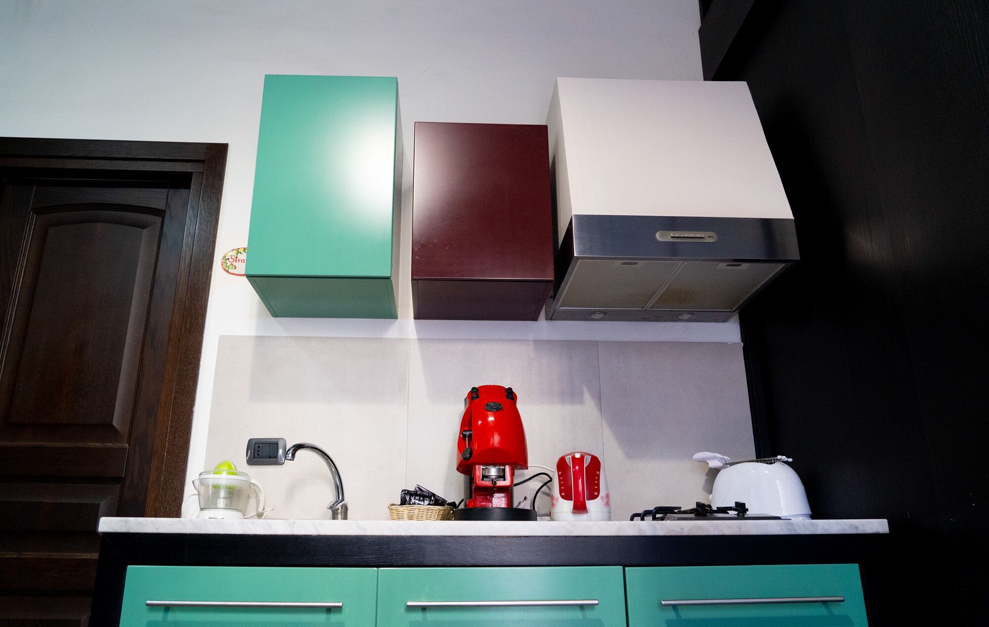 Une cuisine avec un plan de travail blanc, une machine à café rouge, une bouilloire blanche et des placards de couleur turquoise.