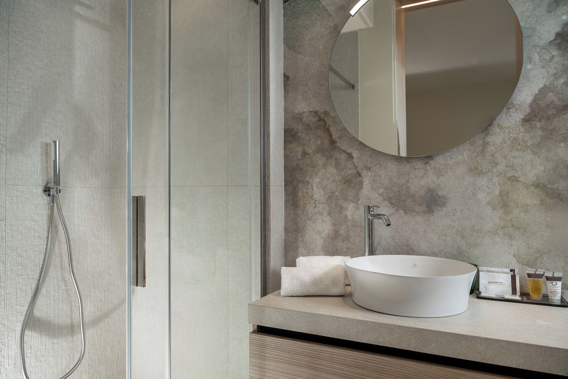 A modern bathroom with a glass shower enclosure, a round mirror, and a white sink with toiletries.