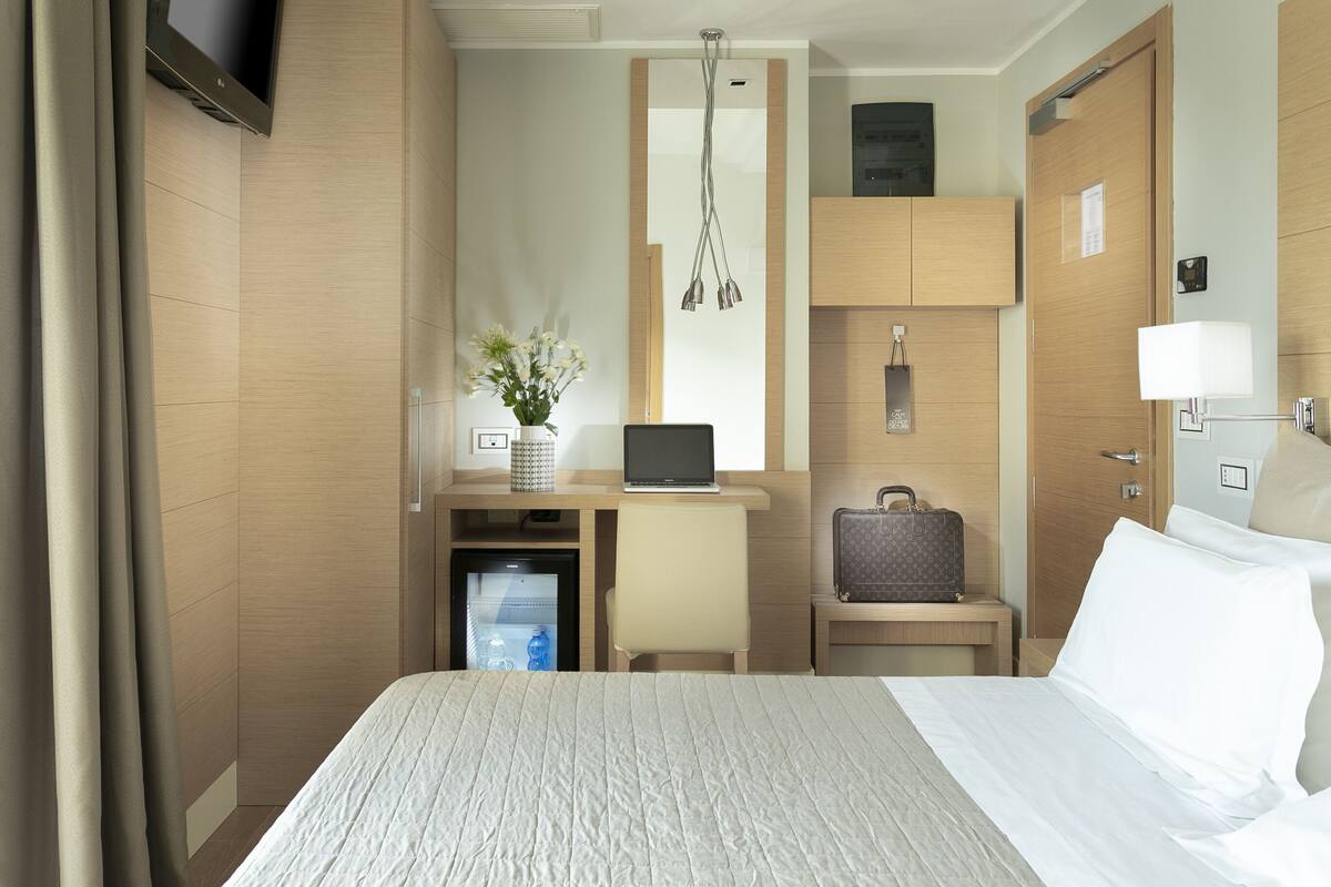 Una habitación de hotel con una cama, un escritorio con una computadora portátil, un refrigerador pequeño, una maleta y un televisor.