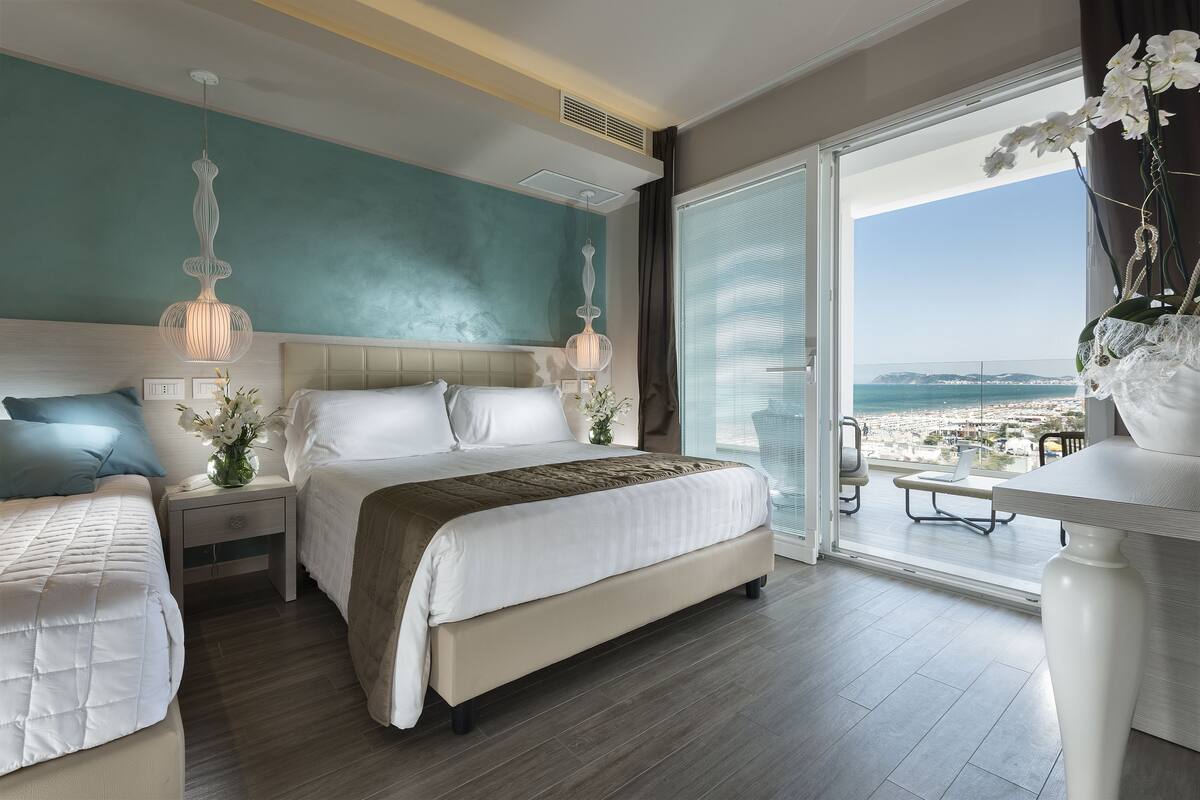 Habitación de hotel con una cama grande, mesitas de noche, un balcón con mobiliario exterior y vistas a la playa.