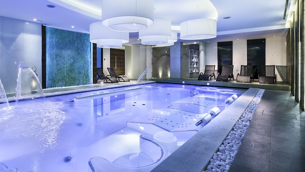 Una piscina interior moderna con agua cristalina, sillas de descanso y una pared grande con diseño texturizado.