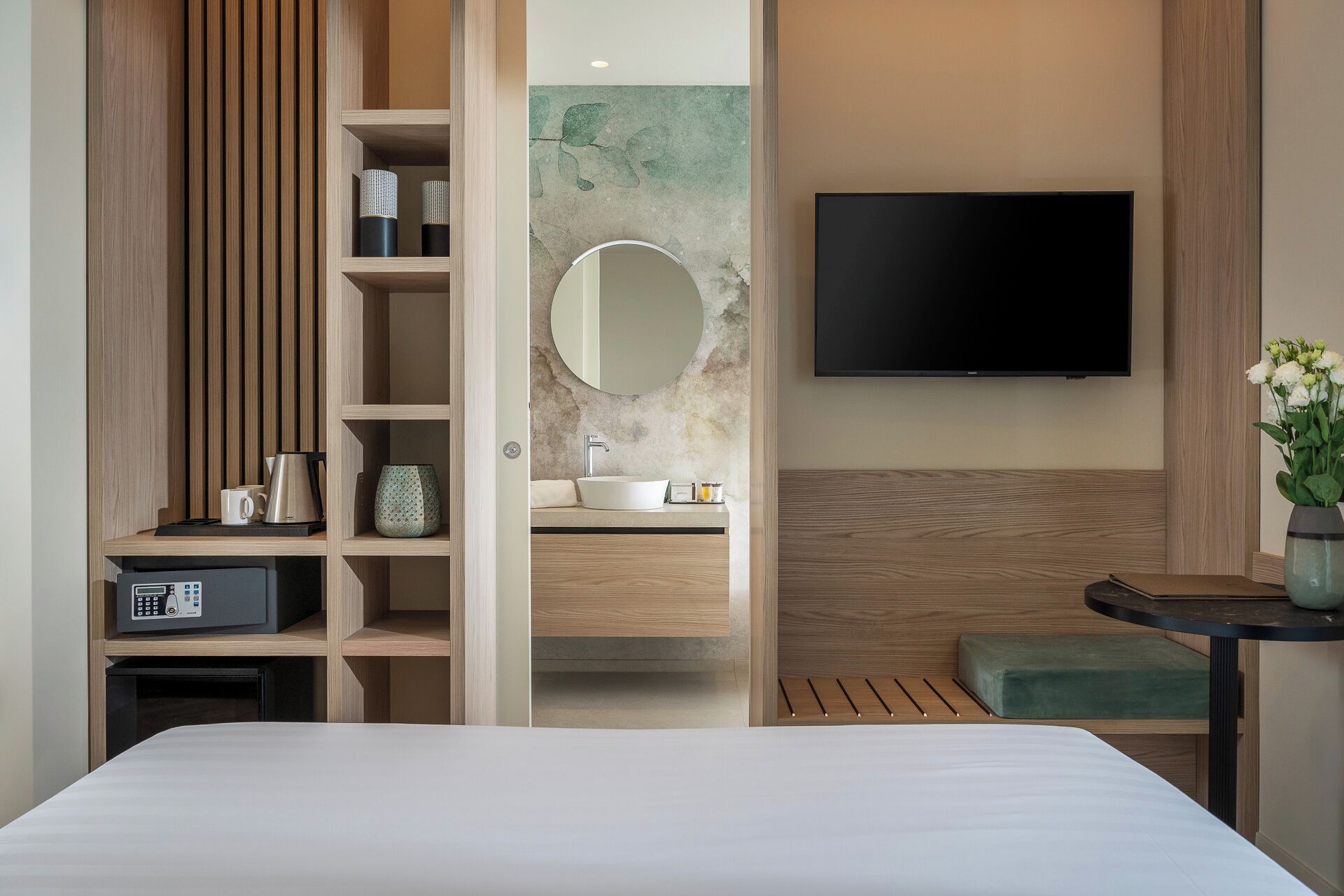 A modern hotel room with a bed, a TV mounted on the wall, a wooden vanity with a sink, and a small seating area.