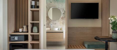 A modern hotel room with a bed, a TV mounted on the wall, a wooden vanity with a sink, and a small seating area.