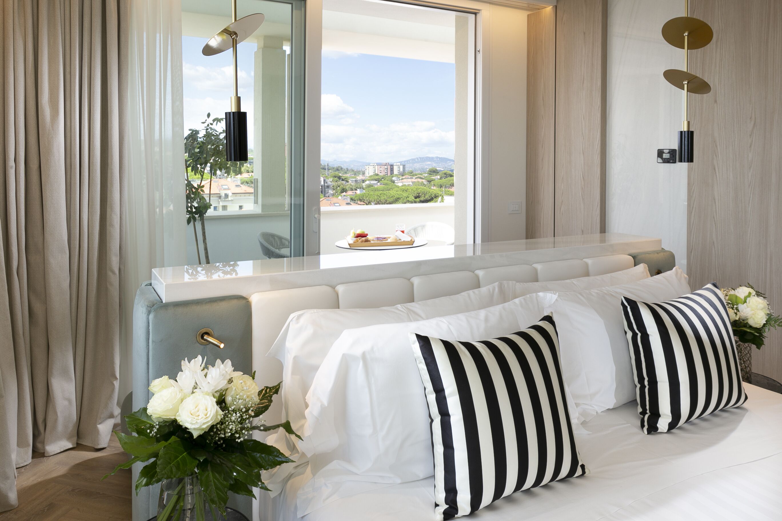 A modern hotel room with a large bed, a view of the city, and a dining area with a tray of food.