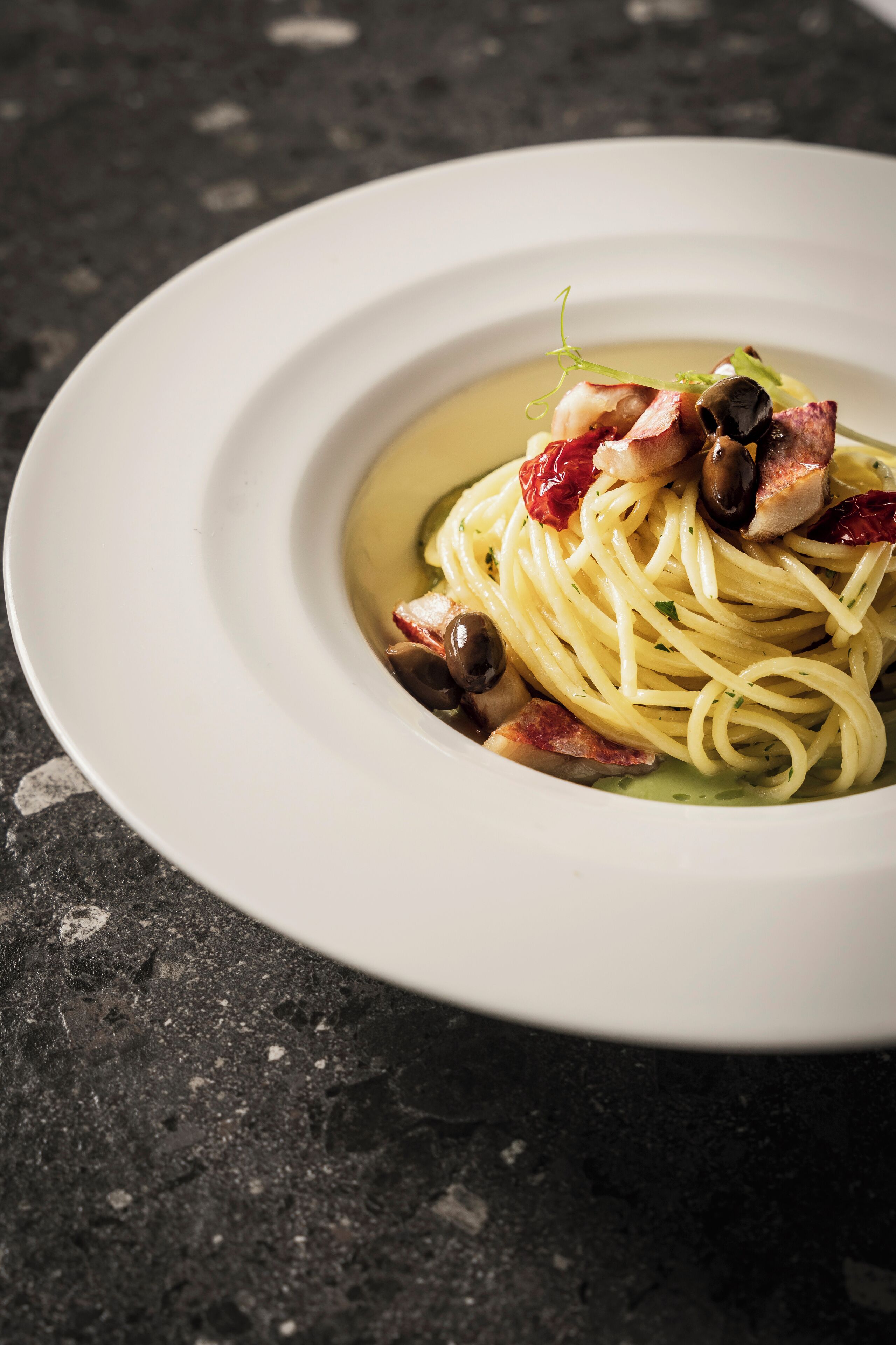 Une assiette blanche contenant des spaghettis, des olives et des lardons, posée sur une surface sombre.